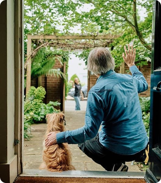 An older man with gray hair, sitting at an open doorway with his dog, waves goodbye to a woman leaving through a garden gate