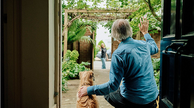 An older man with gray hair, sitting at an open doorway with his dog, waves goodbye to a woman leaving through a garden gate